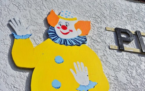 A colorful wooden clown figure with a polka dot hat and blue collar, waving against a textured wall.