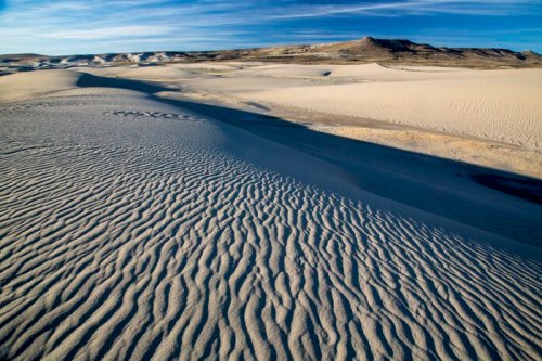 A vast desert landscape with rippled sand dunes under a clear blue sky and distant hills.