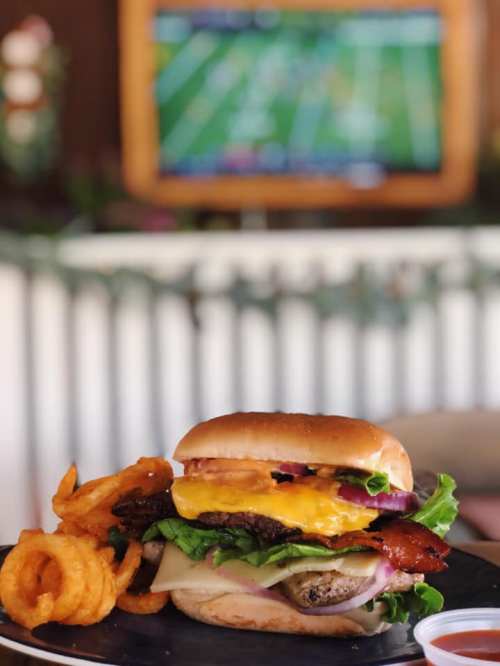A close-up of a burger with lettuce, tomato, cheese, and bacon, served with curly fries, with a football game on TV in the background.
