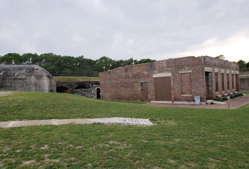 A historic brick building next to a grassy area, with a stone structure in the background under a cloudy sky.