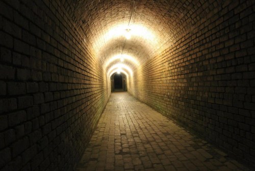 A dimly lit brick tunnel with lights along the ceiling, leading into darkness at the far end.