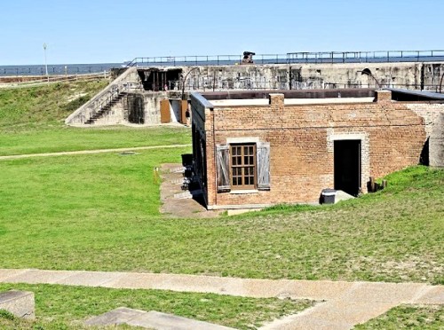 Historic brick building on grassy grounds with a coastal view, surrounded by stone structures and a clear blue sky.