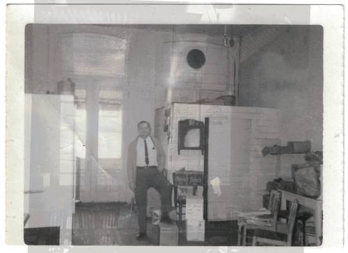 A man in a suit stands beside boxes in a vintage room with large windows and a phone booth.