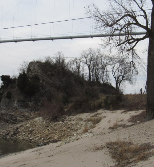 A rocky outcrop near a riverbank, with sparse trees and a power line overhead on a cloudy day.