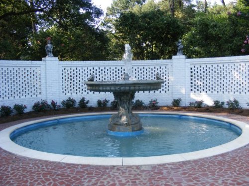A decorative stone fountain in a circular pool, surrounded by a white lattice wall and blooming flowers.