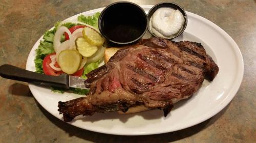 A grilled steak on a plate, accompanied by salad, pickles, tomatoes, and two dipping sauces.