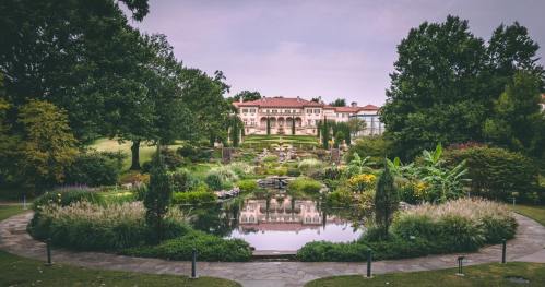 A serene garden with a pond, lush greenery, and a grand building in the background under a cloudy sky.