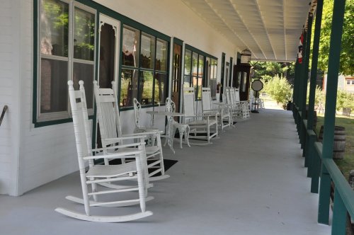 A porch with several white rocking chairs lined up along a green railing, surrounded by trees and a peaceful setting.