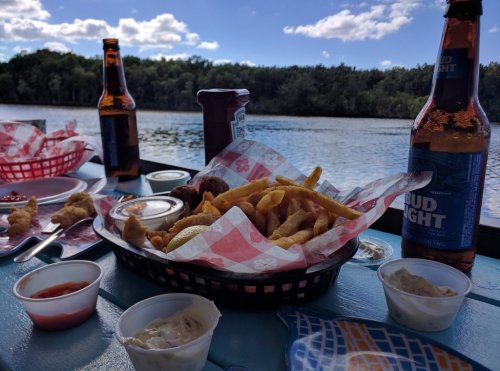 A basket of fried food with dipping sauces, two beer bottles, and a scenic view of water and trees in the background.