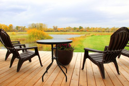 A serene lakeside view from a wooden deck with two chairs and a small table, surrounded by autumn foliage.