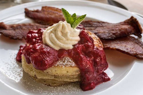 Fluffy pancakes topped with raspberry sauce and whipped cream, served with crispy bacon and a mint garnish.