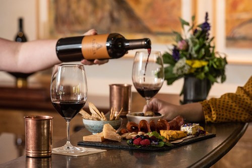 A hand pours red wine into a glass beside a platter of cheese, meats, and fruits, with flowers in the background.