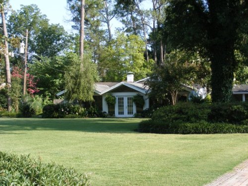 A charming single-story house surrounded by lush greenery and trees, with a well-maintained lawn in front.