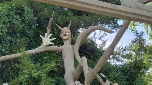 A whimsical sculpture of a creature with a playful expression, made from branches and clay, surrounded by greenery.
