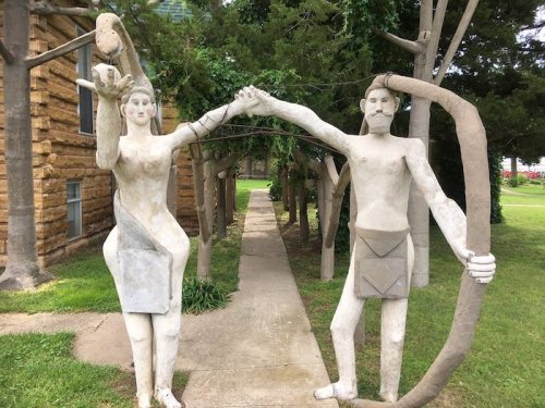 Two abstract sculptures of a man and woman holding hands, set in a grassy area with trees and a pathway.