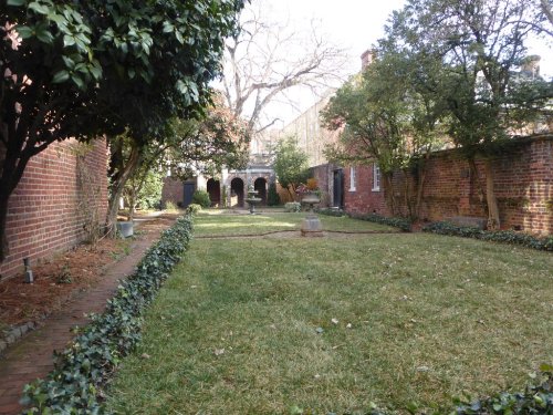 A serene garden with brick walls, green grass, and trees, featuring a pathway leading to an archway in the background.