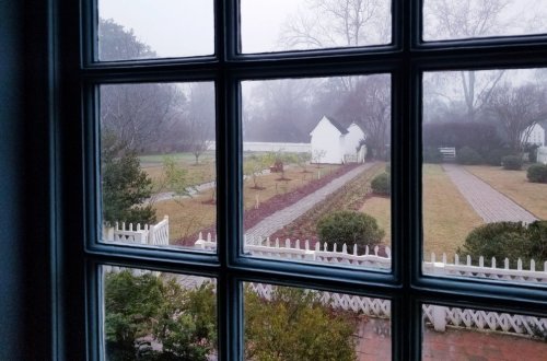 View through a window of a foggy garden with pathways, shrubs, and a white building in the background.