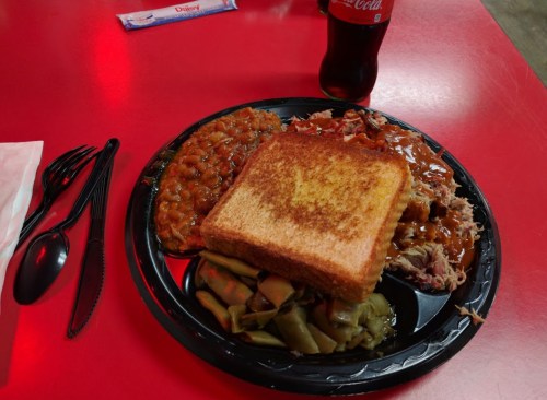 A black plate with pulled pork, baked beans, green beans, toast, and a bottle of Coca-Cola on a red table.