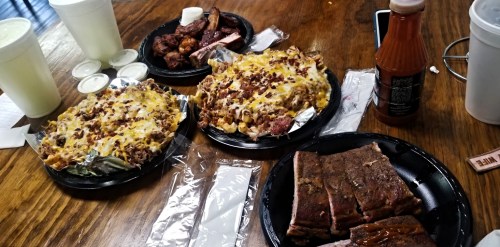 A table with three plates of loaded nachos, BBQ ribs, and wings, accompanied by drinks and sauces.