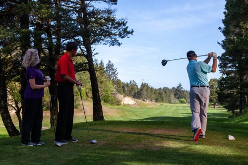 A golfer swings a club on a course, while two spectators watch from the tee box surrounded by trees.