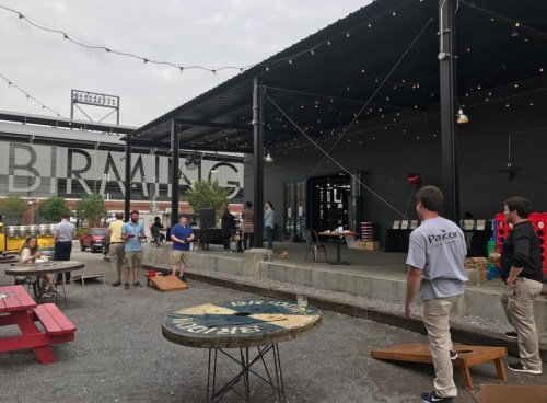 Outdoor gathering with people playing cornhole, string lights overhead, and a large "BIRMINGHAM" sign in the background.