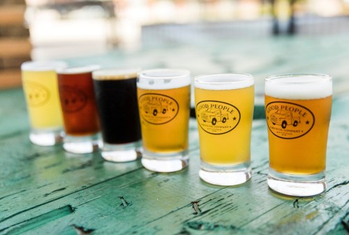 A row of six beer glasses on a wooden table, showcasing various beer styles with a logo on each glass.