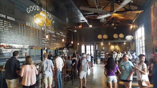 A bustling brewery interior with patrons enjoying drinks, a large menu board, and industrial decor.