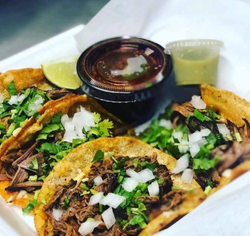 A takeout container with three tacos filled with shredded beef, topped with onions and cilantro, alongside sauces and lime.