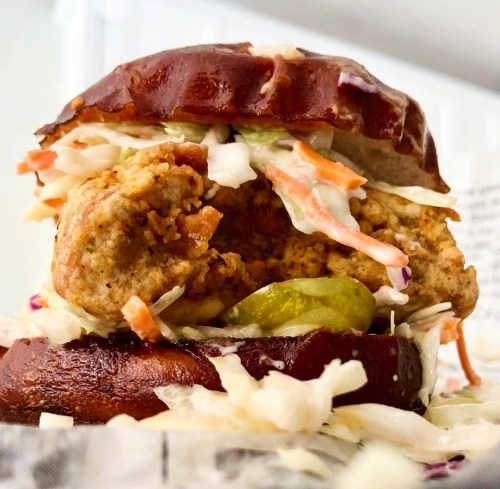 A close-up of a pretzel bun sandwich filled with crispy fried chicken, coleslaw, and pickles.