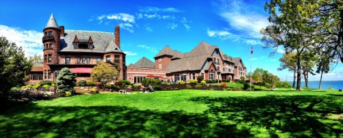 A large, historic mansion with a tower, surrounded by lush green lawns and trees, overlooking a serene lake under a blue sky.