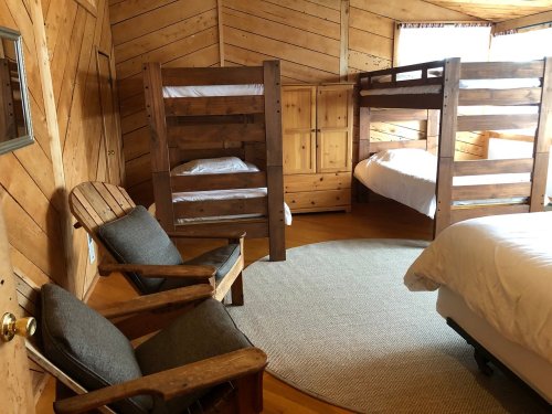 Cozy wooden room with bunk beds, a double bed, and a comfortable chair, featuring warm wood paneling and a round rug.