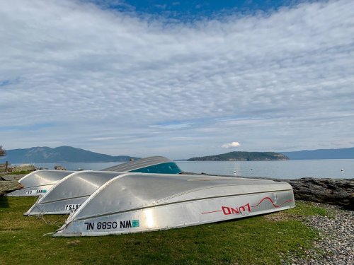 Three silver boats are resting on grass by the water, with a cloudy sky and distant hills in the background.