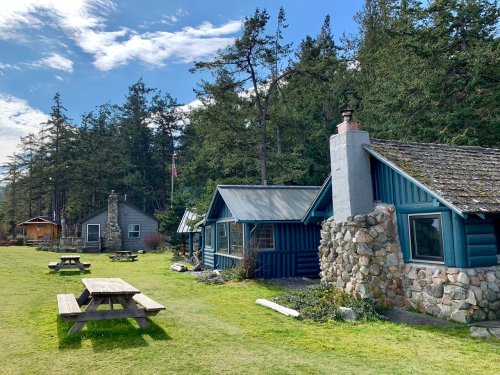 A scenic view of rustic cabins surrounded by trees, with picnic tables on a grassy area under a blue sky.