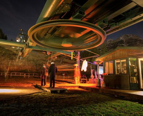 Two people stand near a ski lift at night, illuminated by warm lights, with mountains in the background.
