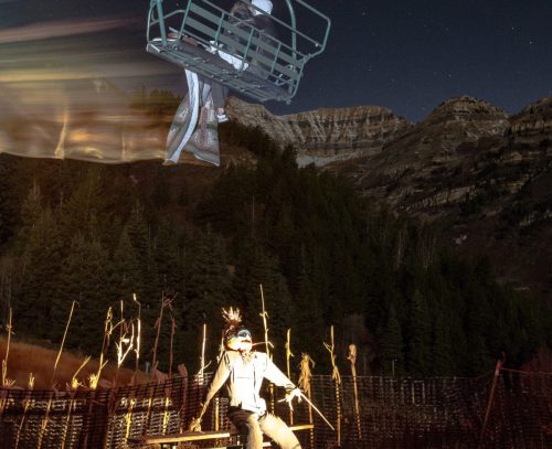 A person sits on a bench at night, with a ski lift overhead and mountains in the background, illuminated by soft light.