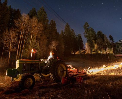 A farmer on a tractor plows a field at night, surrounded by trees and a starry sky.