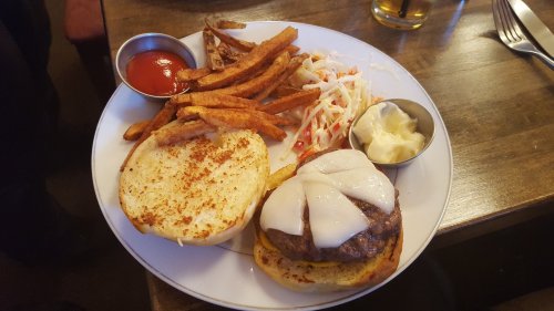 A plate with a cheeseburger, sweet potato fries, coleslaw, and dipping sauces.
