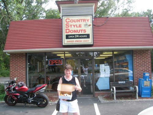 A woman holds a box of donuts outside a donut shop with a red roof and "Open 24 Hours" sign.