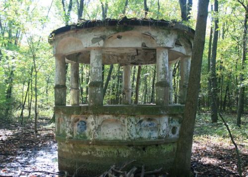 An abandoned, weathered gazebo surrounded by dense green forest, with peeling paint and overgrown vegetation.