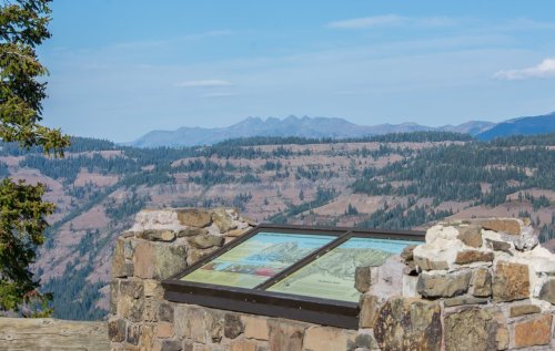 A stone viewpoint with informational panels overlooks a mountainous landscape under a clear blue sky.