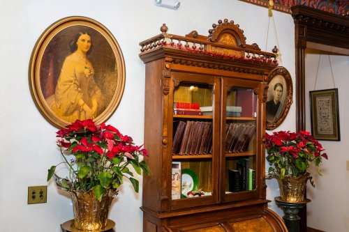 A vintage wooden cabinet with books, surrounded by potted poinsettias and framed portraits on the wall.
