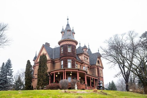 A large, ornate Victorian mansion with a turret, surrounded by trees and a grassy lawn on a cloudy day.