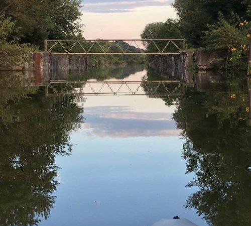A tranquil canal scene with a bridge reflected in the water, surrounded by trees and a soft sky.