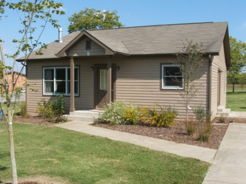 A small, single-story house with a brown exterior, front porch, and landscaped yard on a sunny day.
