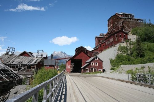 Abandoned red buildings on a gravel road, surrounded by greenery and mountains under a blue sky.