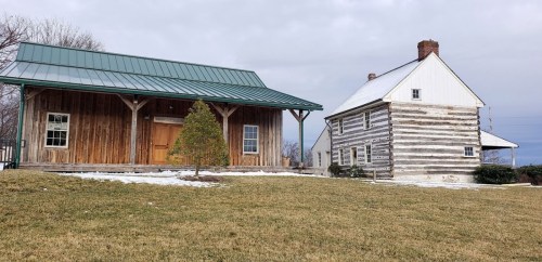 Two rustic buildings: a wooden cabin with a green roof and a log house, set on a grassy area with some snow.