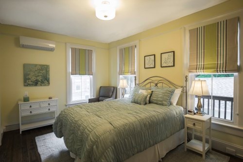 Cozy bedroom with a queen bed, light green bedding, yellow walls, and natural light from two windows with striped shades.