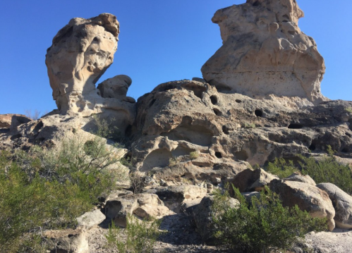 Two large, weathered rock formations against a clear blue sky, surrounded by sparse vegetation.