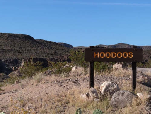 Sign reading "HOODOOS" in a desert landscape with rocky hills and sparse vegetation. Clear blue sky above.