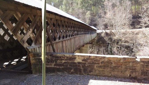 A wooden covered bridge spans a rocky ravine, surrounded by trees and a gravel path.